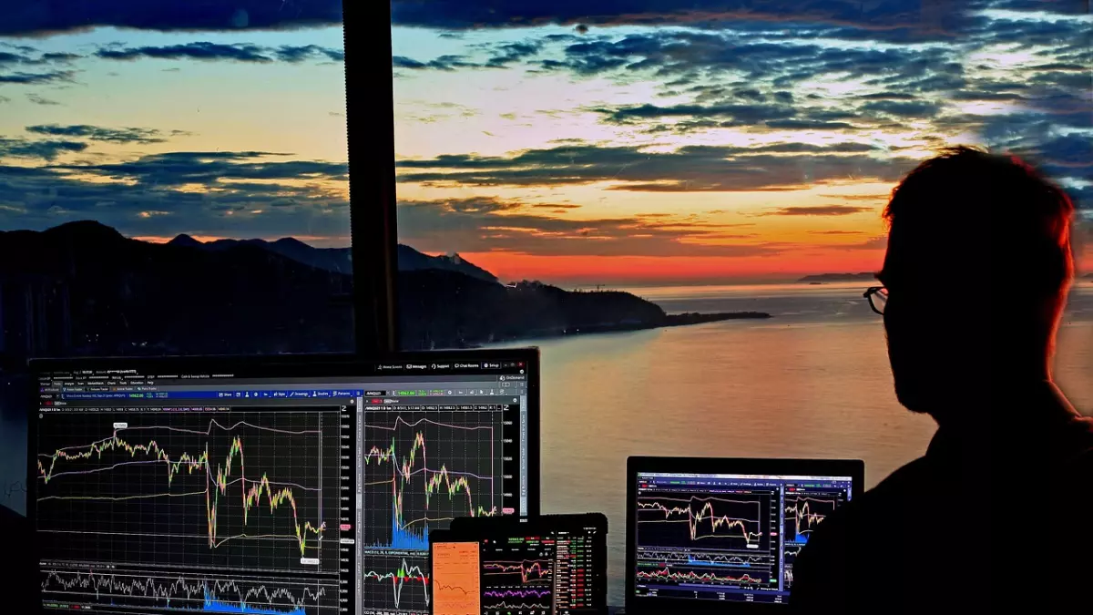 A person sits in front of multiple screens, with a computer monitor, laptop, and tablet. They are all showing stock charts. There is a window behind them that shows a sunset over water.