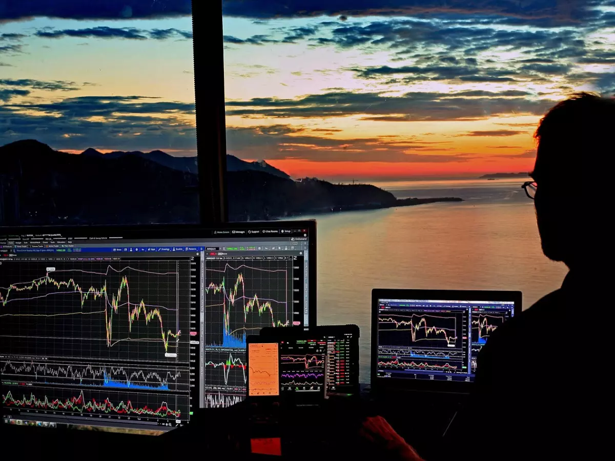 A person sits in front of multiple screens, with a computer monitor, laptop, and tablet. They are all showing stock charts. There is a window behind them that shows a sunset over water.