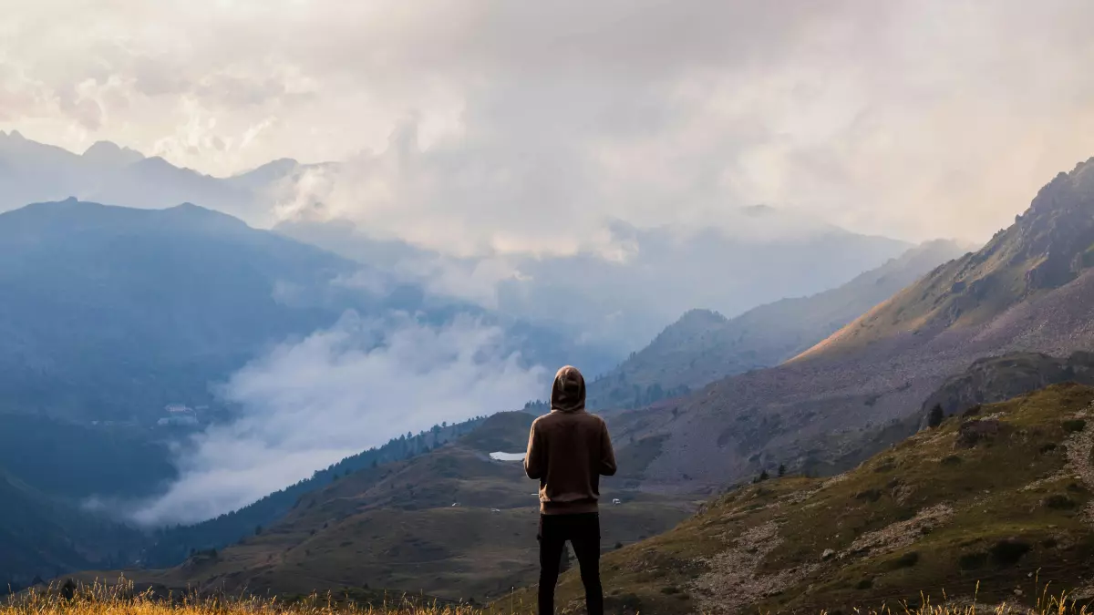 A man stands on a grassy hill, looking out at a vast, mountainous landscape. The mountains are covered in clouds, and the sky is blue.