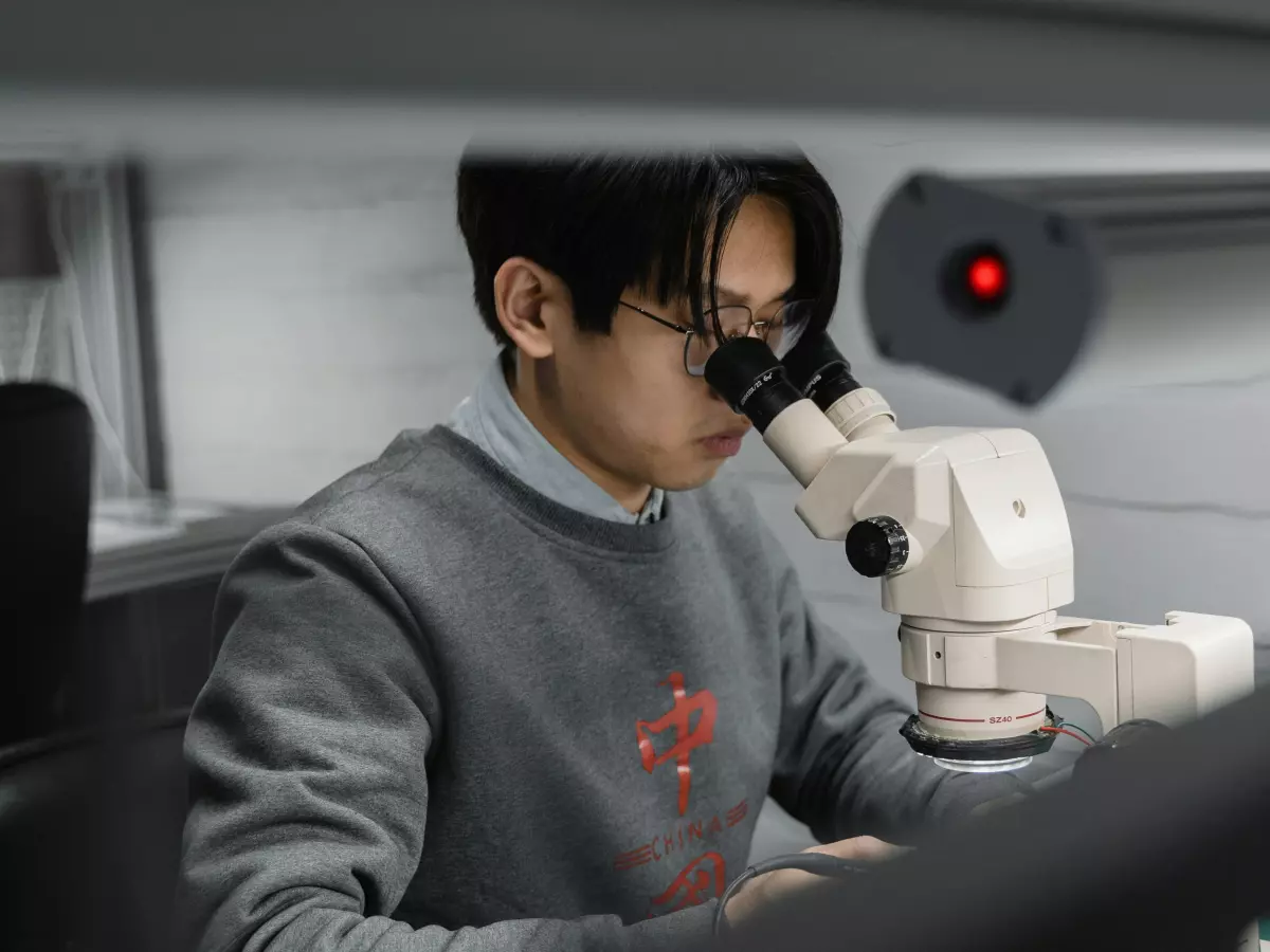 A person is using a microscope to repair a small object. The person is wearing a light blue sweater and glasses. The background is blurred and has a red light.