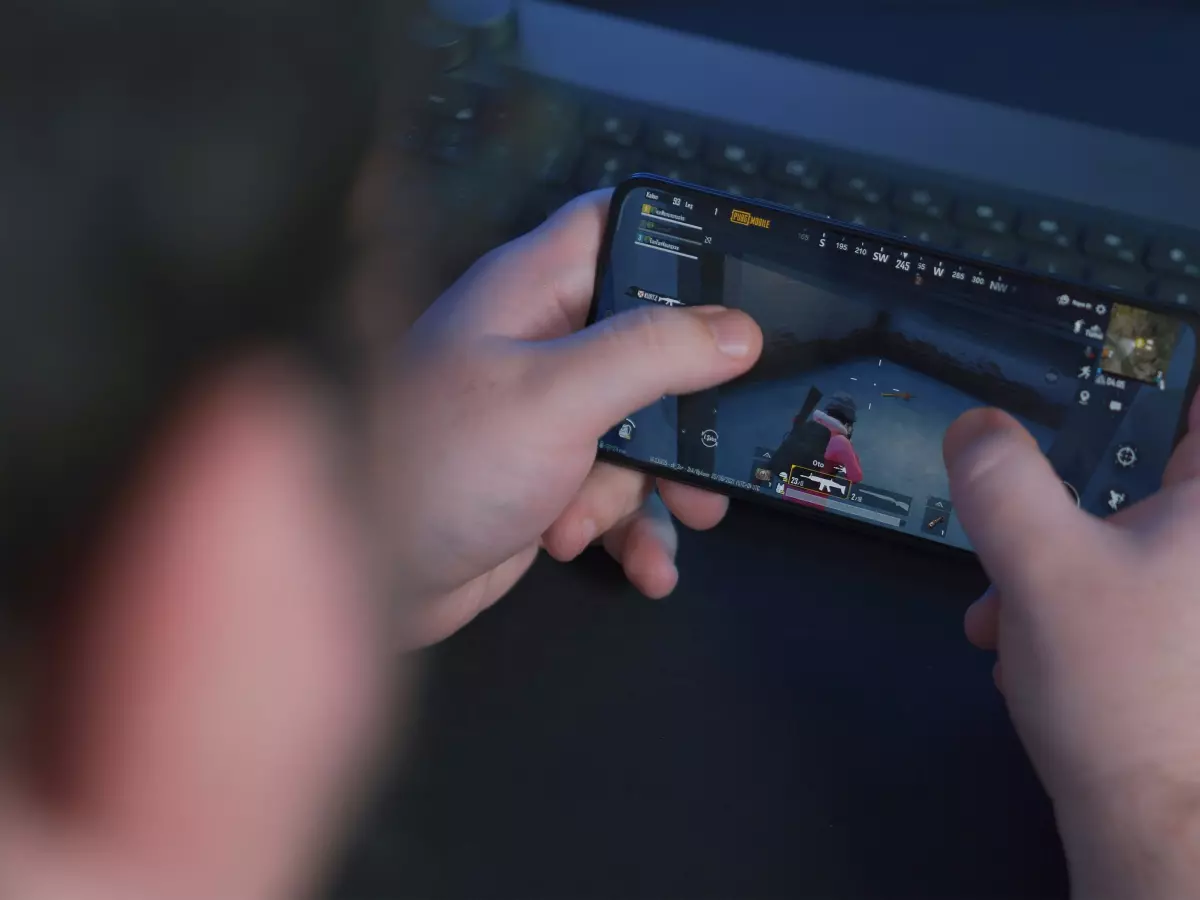 A person is playing a game on their phone while sitting at a desk. The phone is in the center of the image, and the game is a first-person shooter with a dark and gritty visual style. The person is focused on the game, and the background is blurry. There is a keyboard and a pair of glasses on the desk.