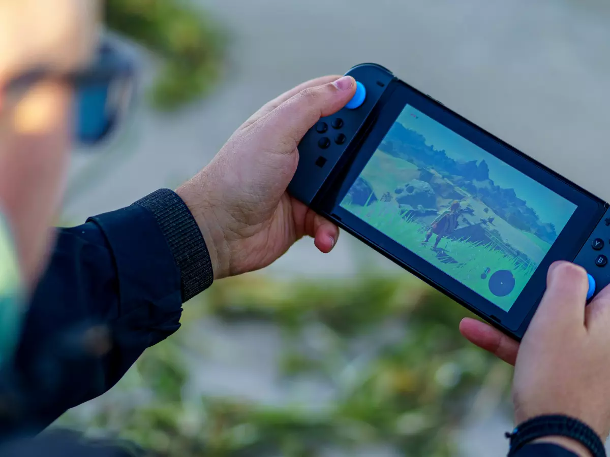 A person is playing a game on a Nintendo Switch handheld console. The person's fingers are on the buttons and joysticks, and the screen shows a colorful landscape with mountains and a lake.