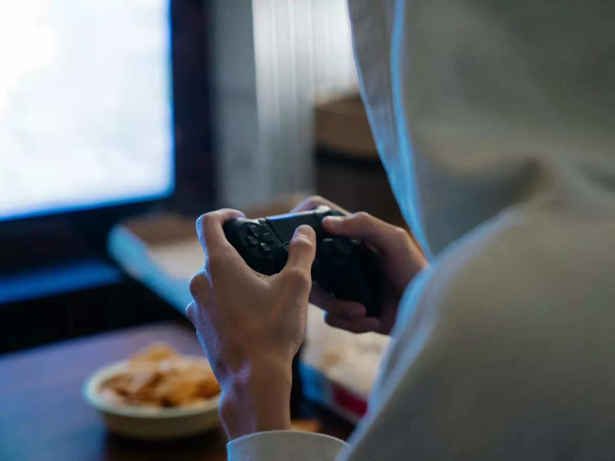 A person in a hooded sweatshirt is playing a game on a handheld device. The person is in a dimly lit room and their face is obscured by the hood.