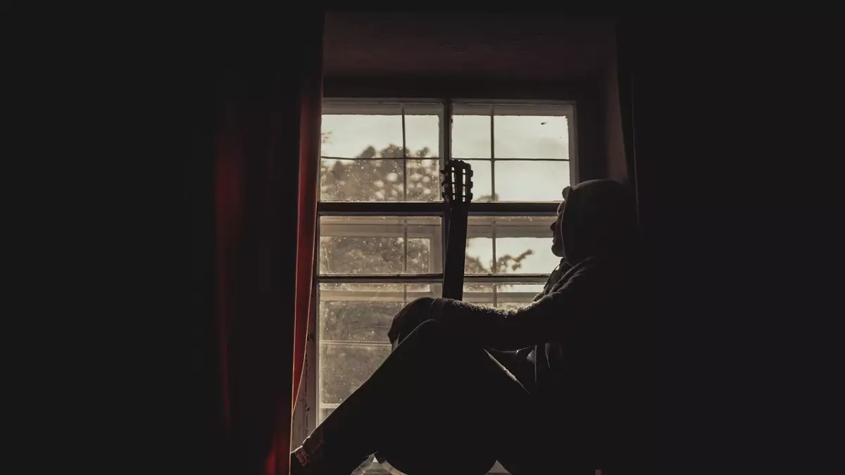 A silhouette of a person sitting by a window with a guitar, looking out at the view.