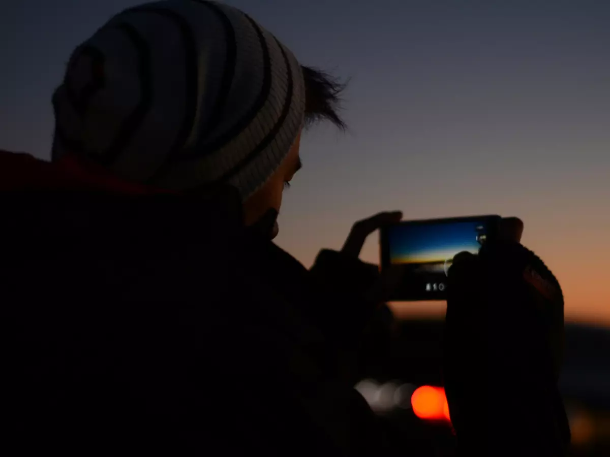 A person silhouetted against a sunset, taking a photo with their smartphone.