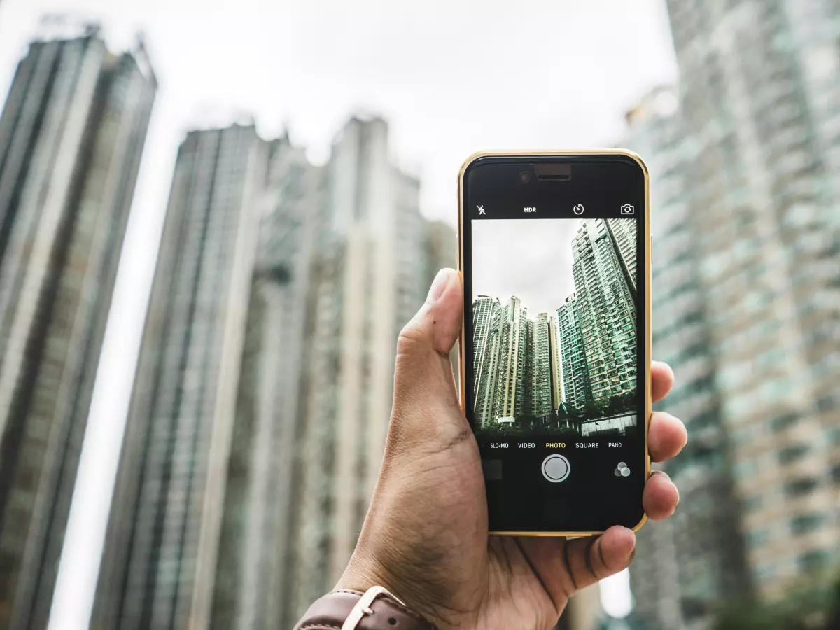 A hand is holding a black smartphone with a gold frame. The phone is facing towards the camera and the screen is displaying a photo of tall buildings. The buildings are in the background and out of focus. The hand is wearing a watch.  The image is taken in the day time, with a cloudy sky and no sunlight