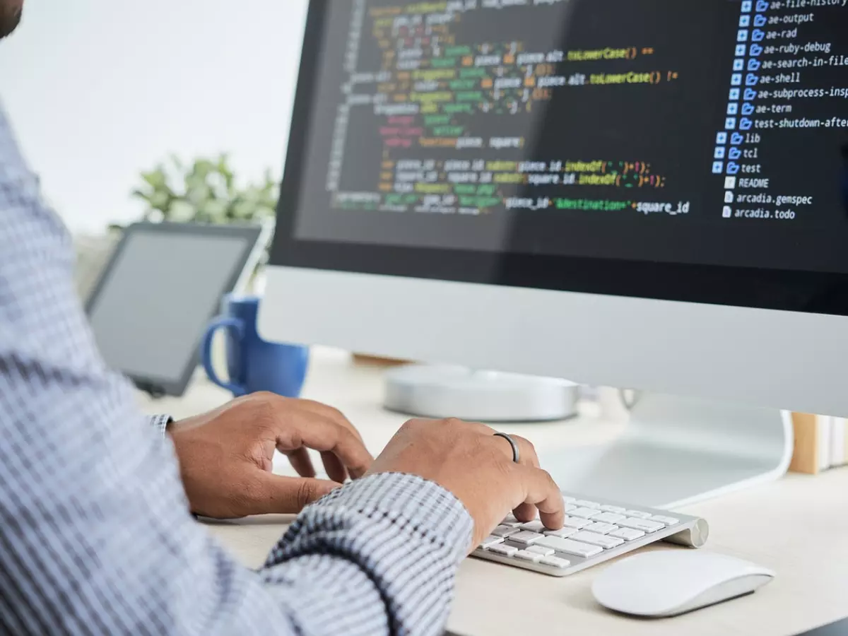 A person's hands are typing on a keyboard with a computer screen showing code in the background.