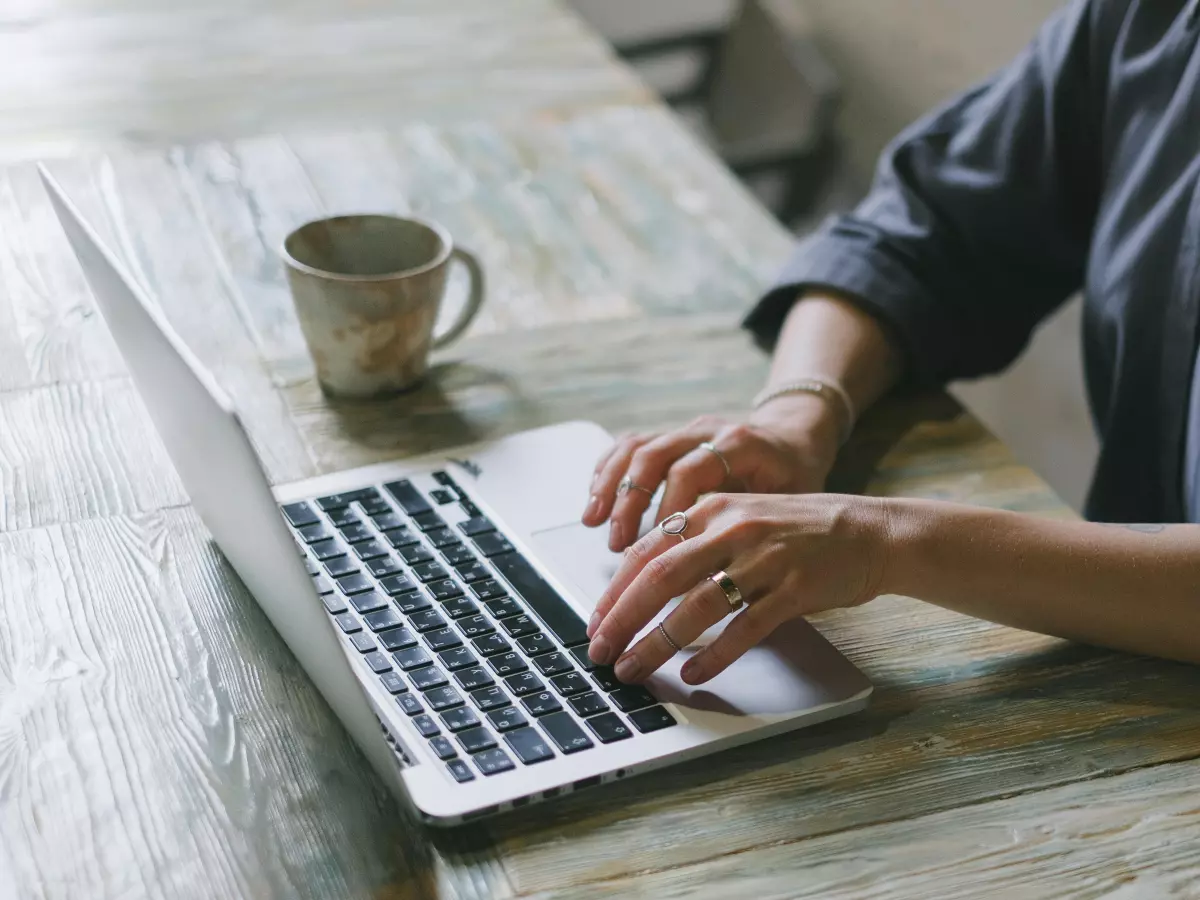 A woman is sitting at a table and typing on a laptop. She is wearing a blue shirt and has her hair pulled back. The table is made of wood and there is a cup of coffee on the table next to the laptop.