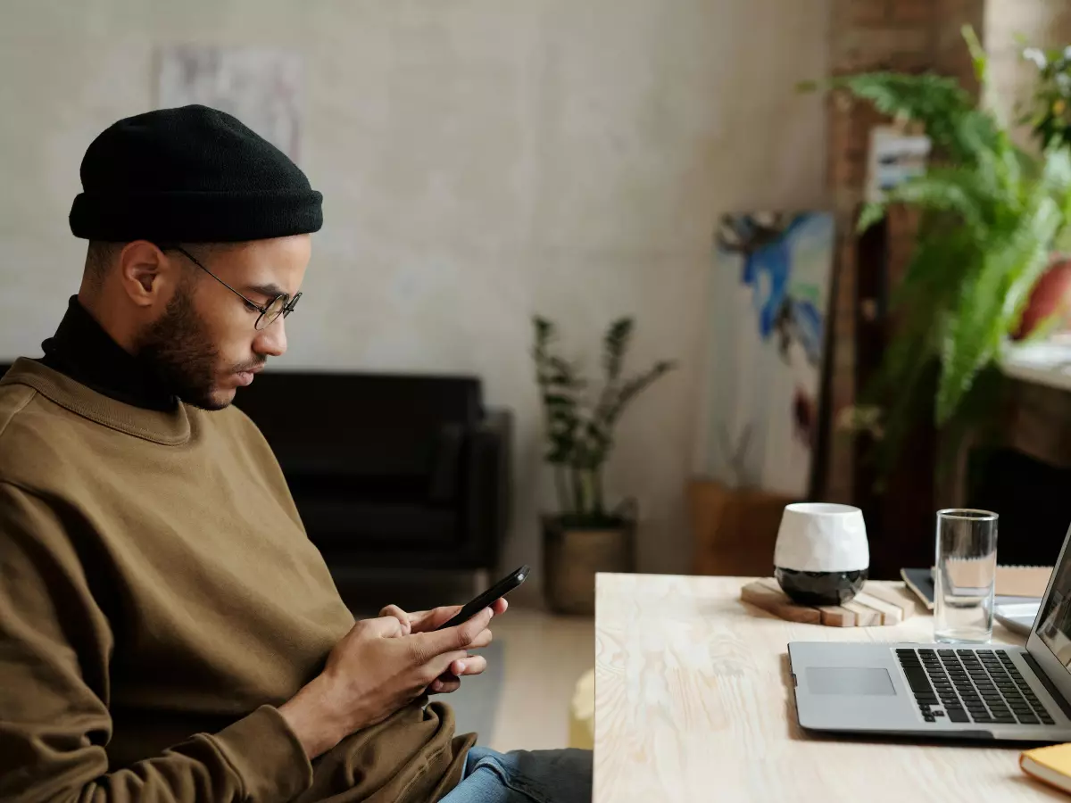 A man is using his smartphone while sitting at a desk. He is wearing a brown sweater and a black beanie. There is a laptop and a plant in the background. 