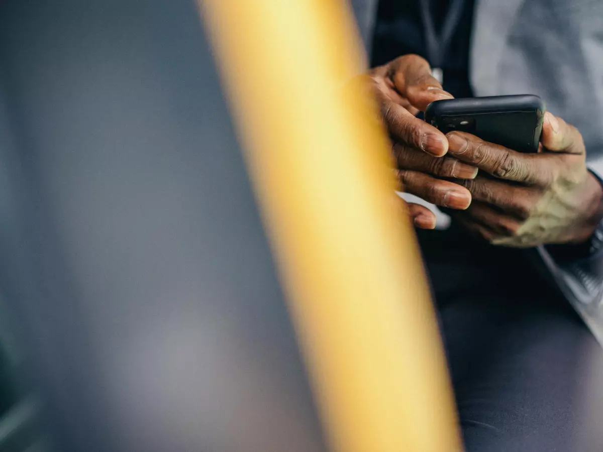 Close-up of a person's hands holding a smartphone in a car. The background is blurred, and the focus is on the hands and the phone. The person is wearing a suit and is sitting in the passenger seat.