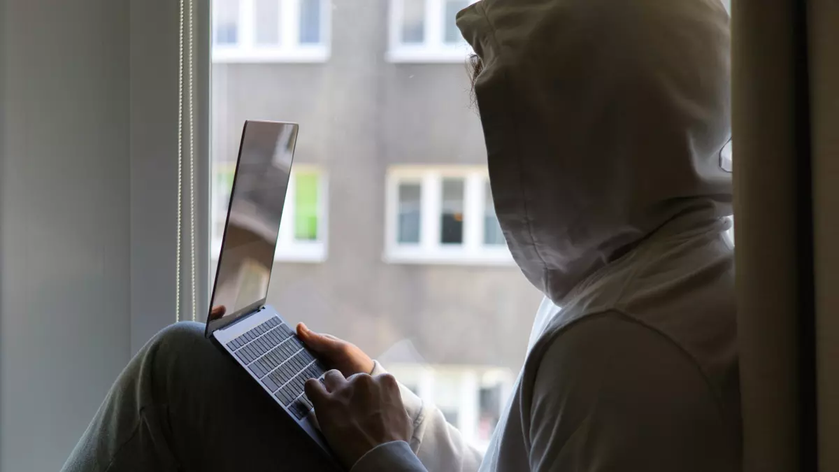 A person in a hoodie is sitting by a window, using a laptop.