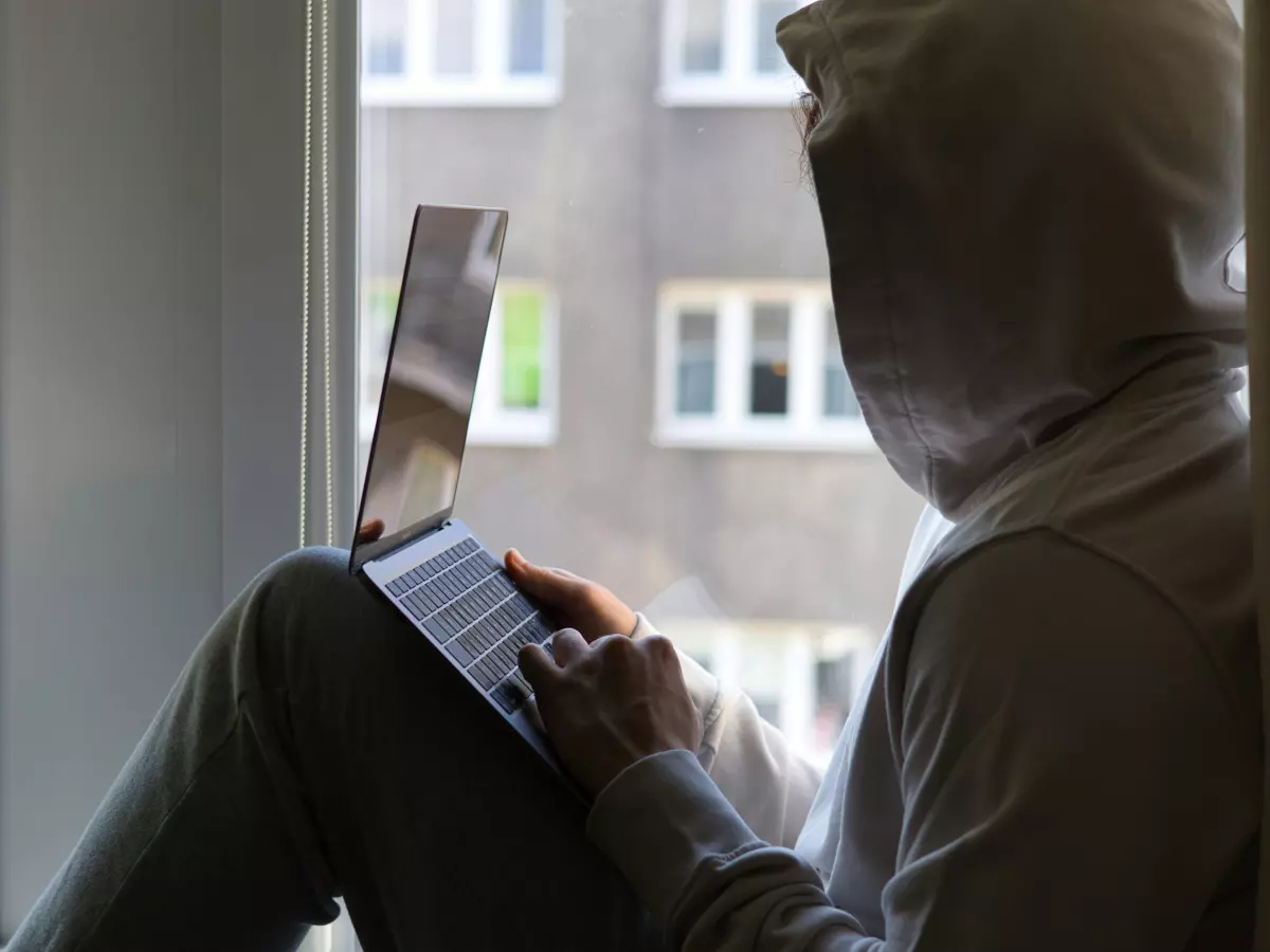 A person in a hoodie is sitting by a window, using a laptop.