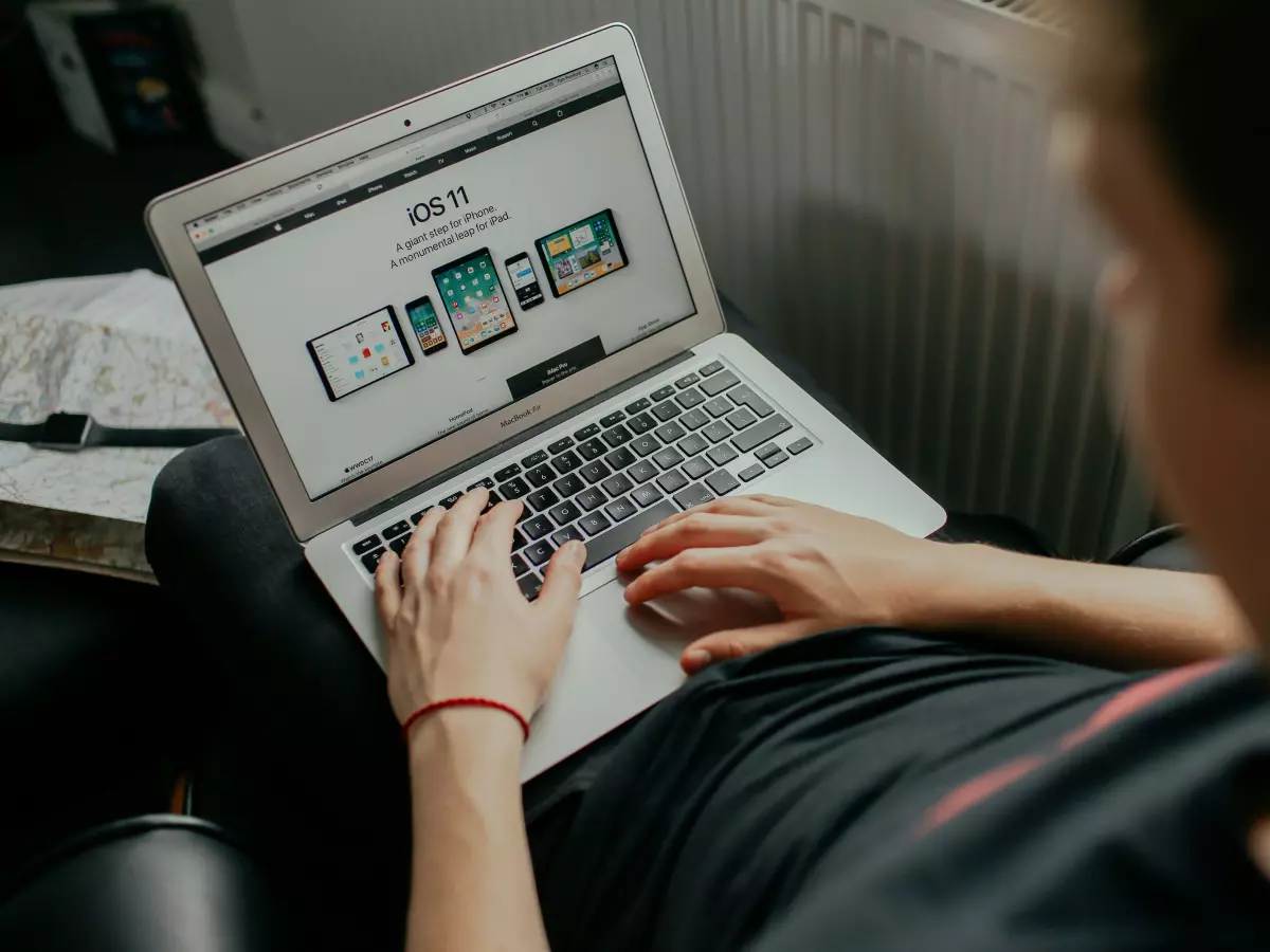 A person sitting on a couch, working on a laptop. The person is wearing a grey hoodie, and is looking at the screen. The laptop is on their lap, and they are using their left hand to type.