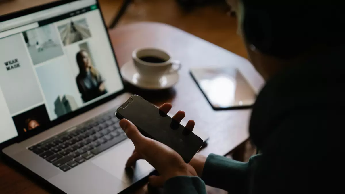 A person holding a smartphone in front of a laptop, with a cup of coffee in the background.