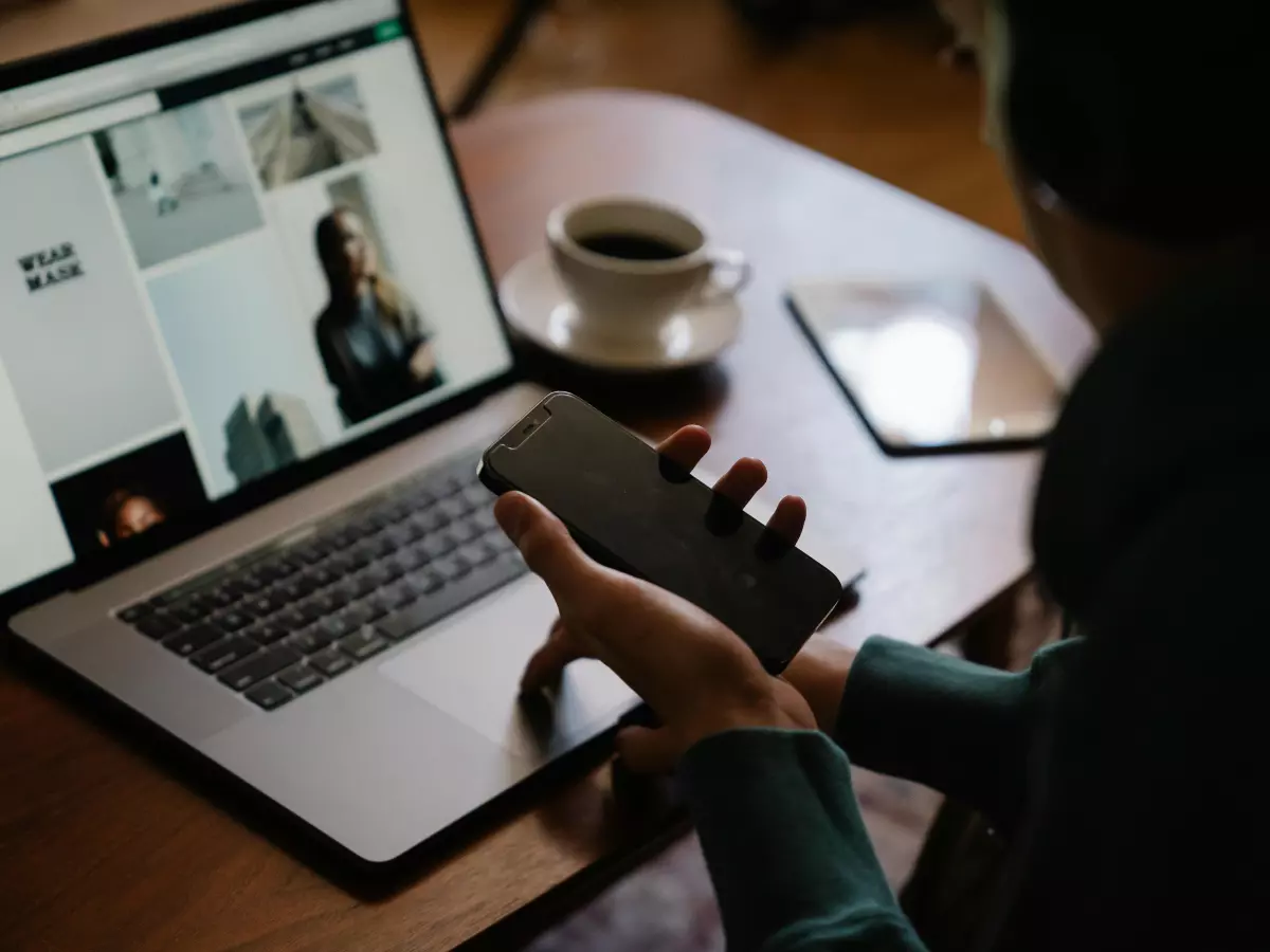 A person holding a smartphone in front of a laptop, with a cup of coffee in the background.