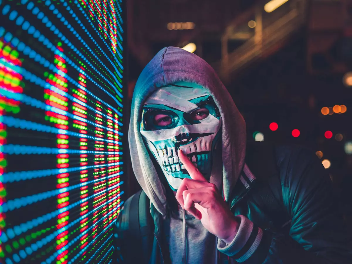 A person wearing a skull mask, with their finger to their mouth, standing in front of a wall of glowing digital code.