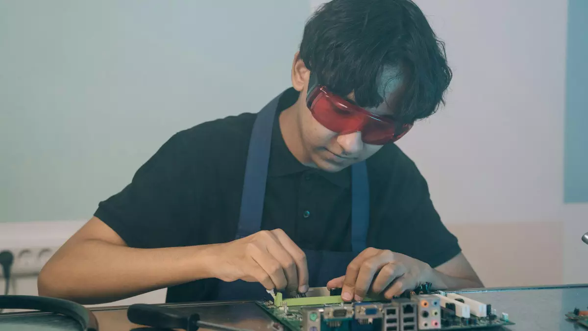 A young man in safety glasses is soldering a circuit board. The image has a blueish hue.