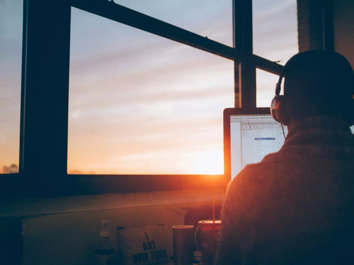 A man is sitting at a computer, wearing headphones, with a sunset view outside the window.