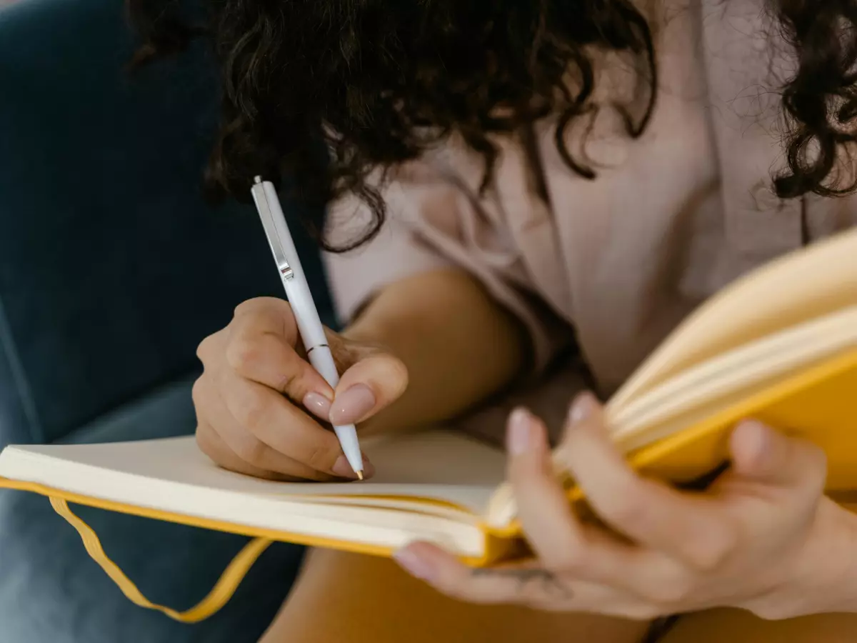 A woman with long, curly hair is sitting on a couch and writing in a notebook. She is wearing a light brown shirt and has a laptop, a cup of coffee, and a smartphone on the table in front of her.