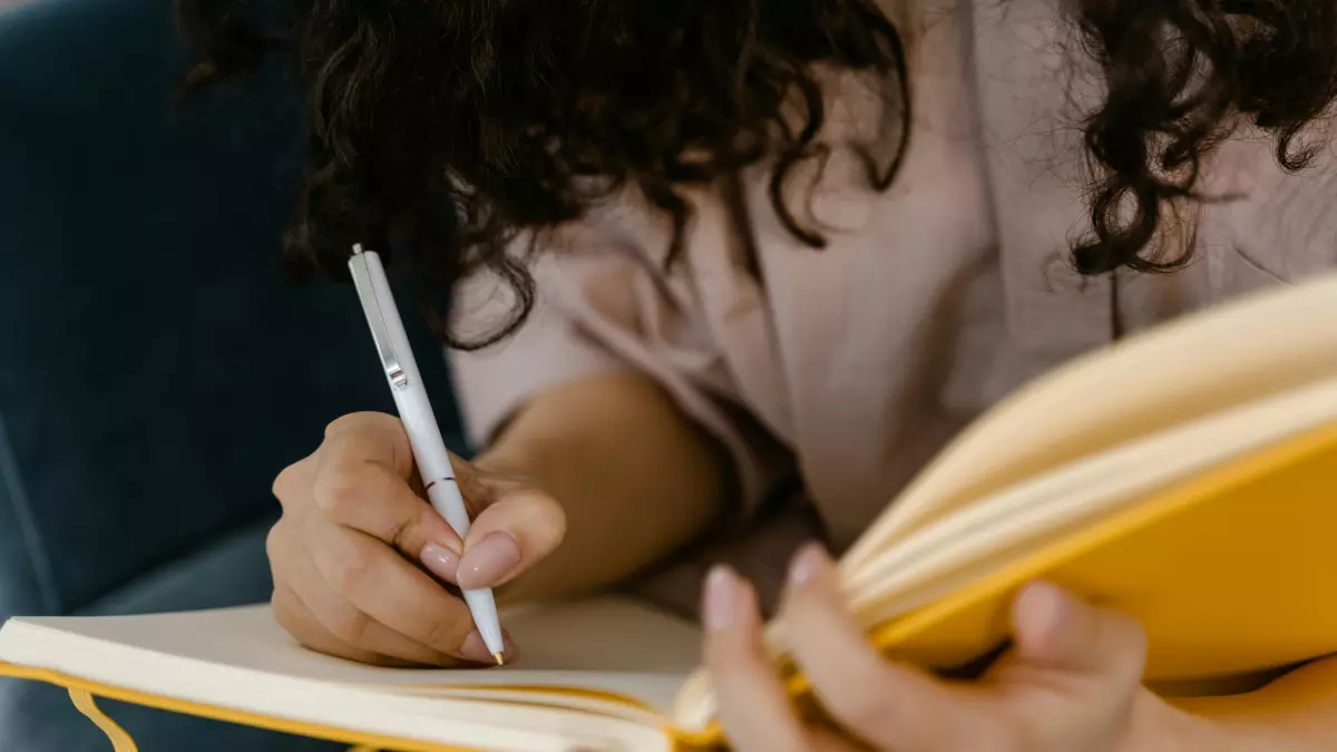 A woman with long, curly hair is sitting on a couch and writing in a notebook. She is wearing a light brown shirt and has a laptop, a cup of coffee, and a smartphone on the table in front of her.