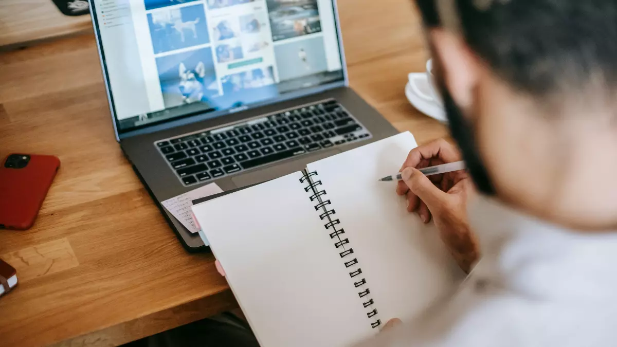 A person is working on a laptop, writing in a notebook.