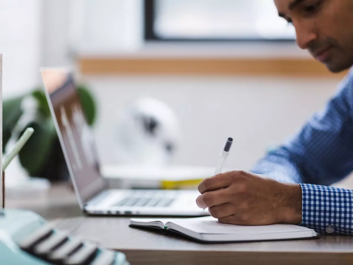 A person's hand writing in a notebook with a laptop in the background.