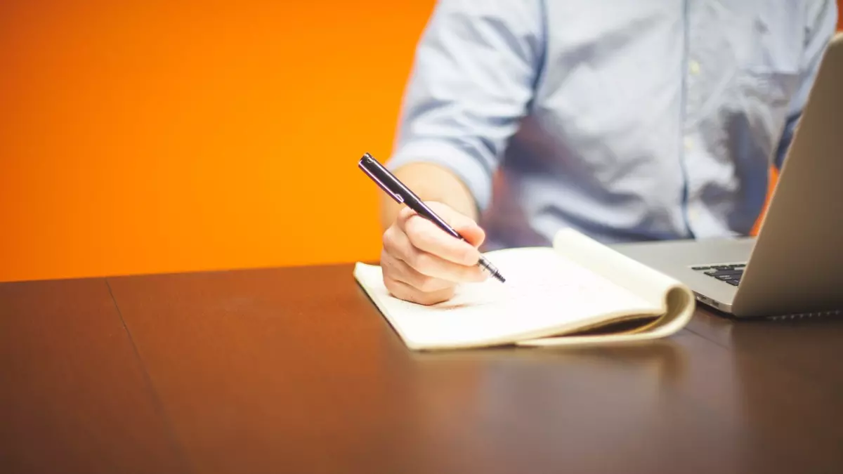 A person is sitting at a desk, writing in a notebook with a pen. A laptop computer is on the table next to them. The background is a bright orange wall. 