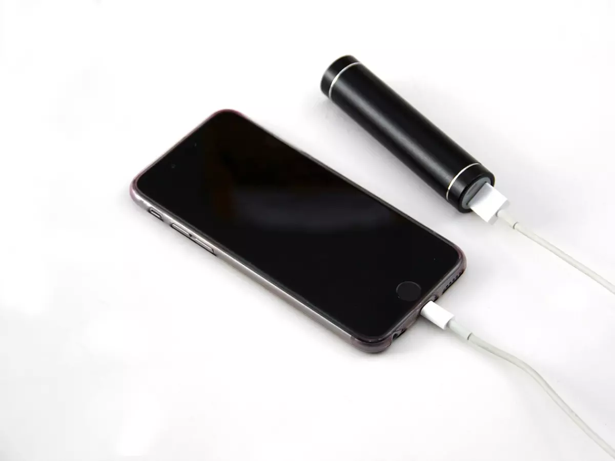 An iPhone is charging with a black power bank. It is connected by a lightning cable. The phone is lying face-down on a white surface.