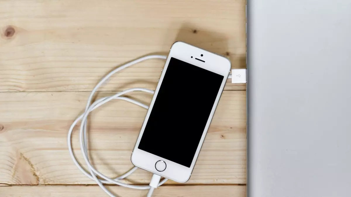 A white iPhone is plugged into a laptop via a white USB cable, lying on a wooden surface. The phone