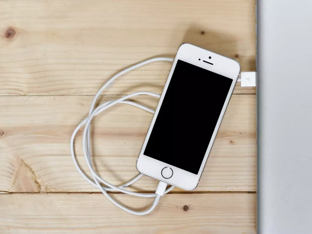 A white iPhone is plugged into a laptop via a white USB cable, lying on a wooden surface. The phone's screen is black.