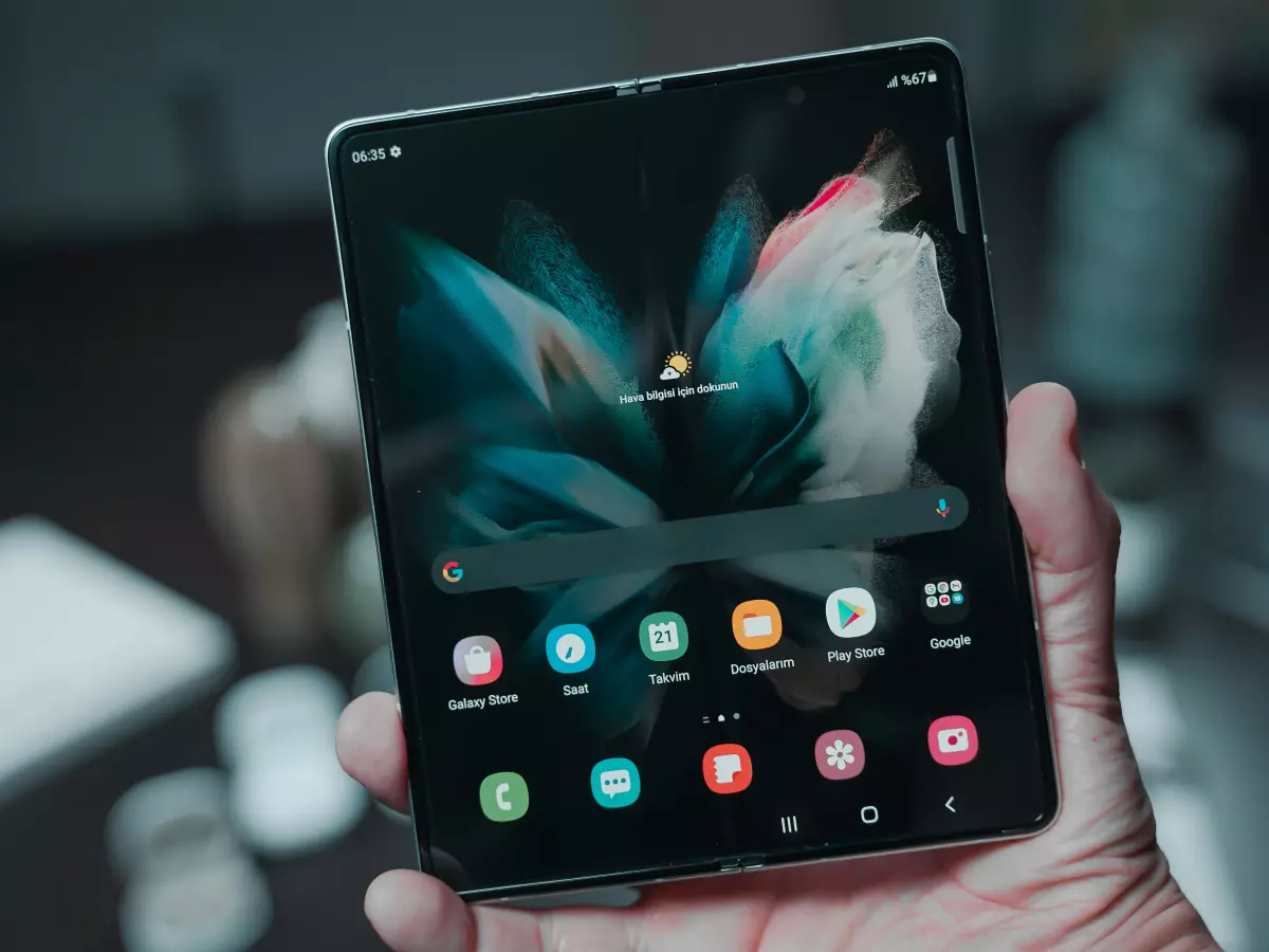 A person holding a Samsung Galaxy Z Fold 6, the phone is unfolded and the screen is displaying the home screen. The phone is in a close-up and the background is blurred.