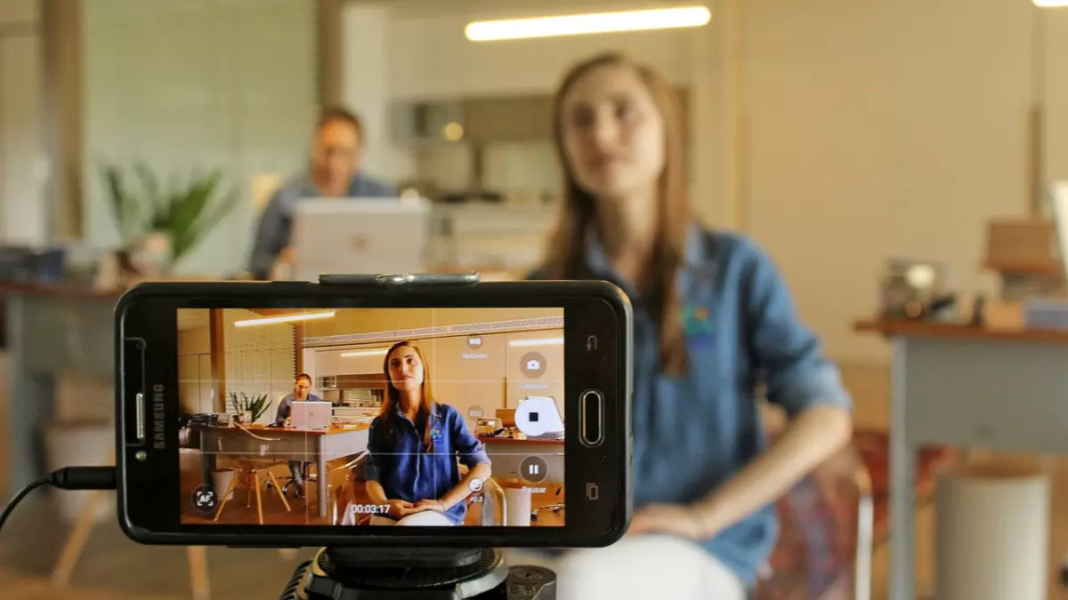 A smartphone is mounted on a tripod and recording a young woman in an office setting. There are two other people in the background.