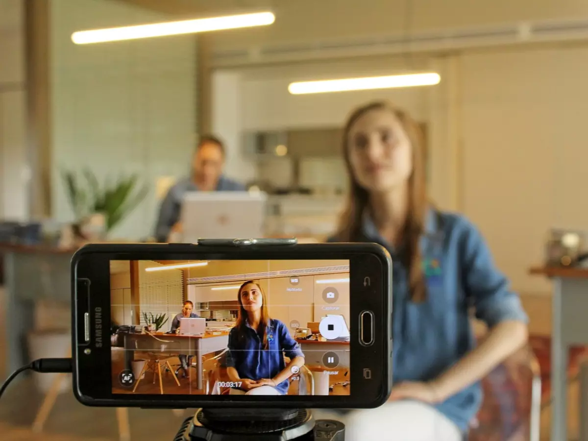 A smartphone is mounted on a tripod and recording a young woman in an office setting. There are two other people in the background.