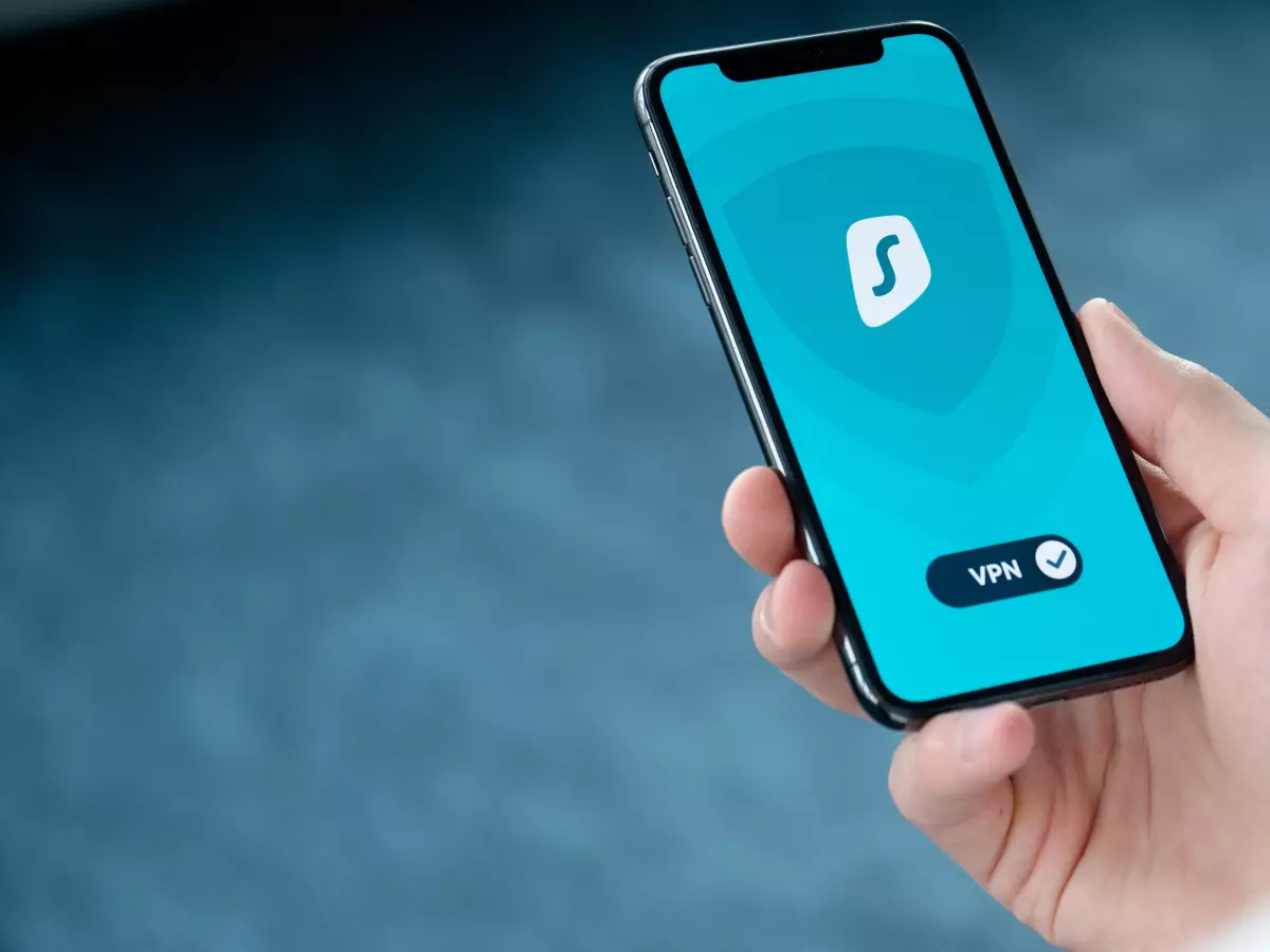 A person holding a smartphone with a security app open, the app icon shows a shield and the word "VPN" is visible, the screen is showing a green check mark indicating that the VPN is connected.