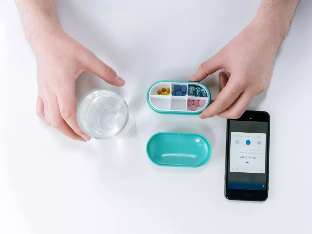 Close-up of a person using an Android phone on a white table, with a pill box and a glass of water in the background.