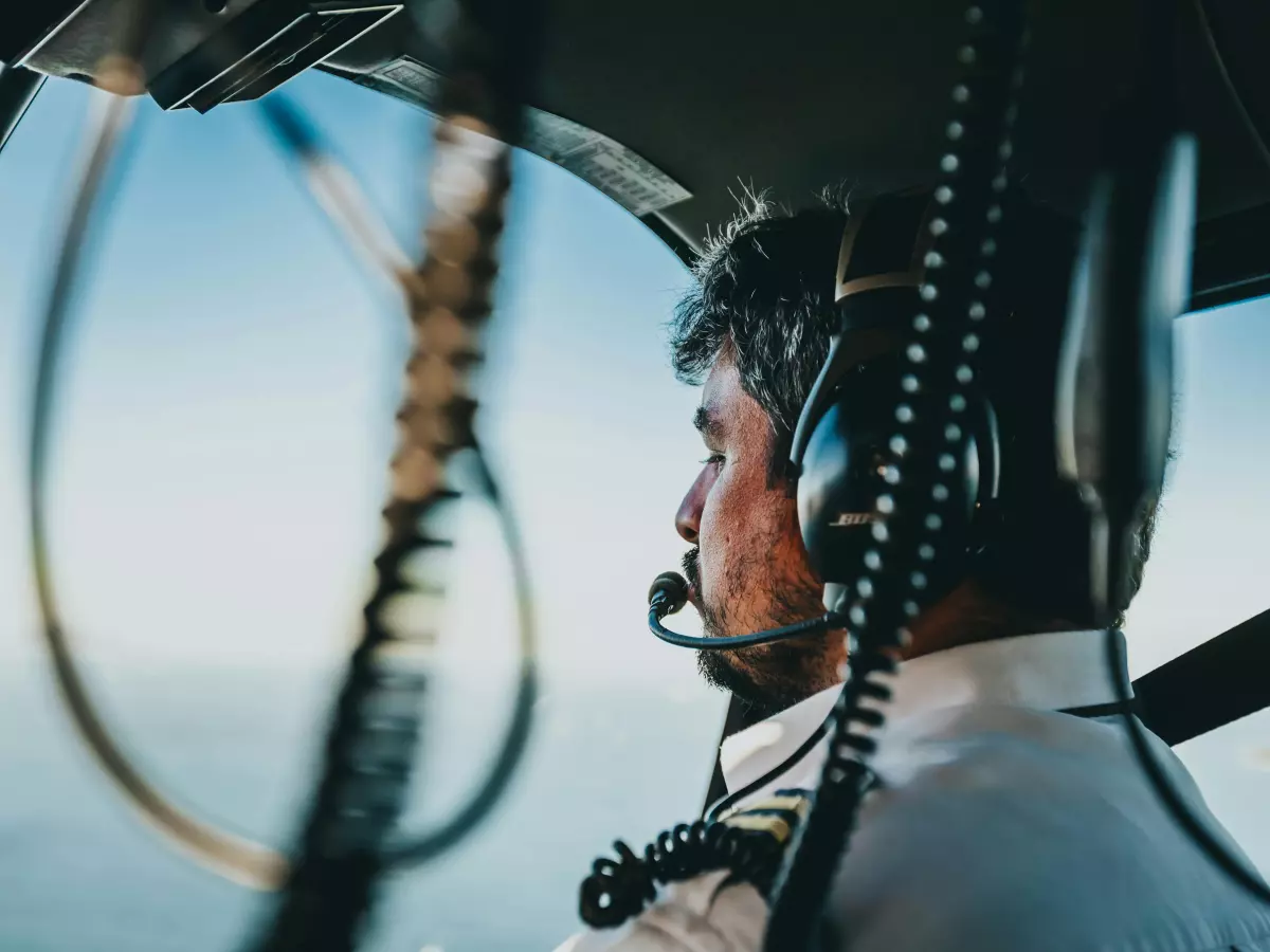 A close-up shot of a pilot in a cockpit, looking out the window, wearing headphones and a microphone. The image is focused on the pilot's face and the headphones, with the rest of the cockpit blurred out.