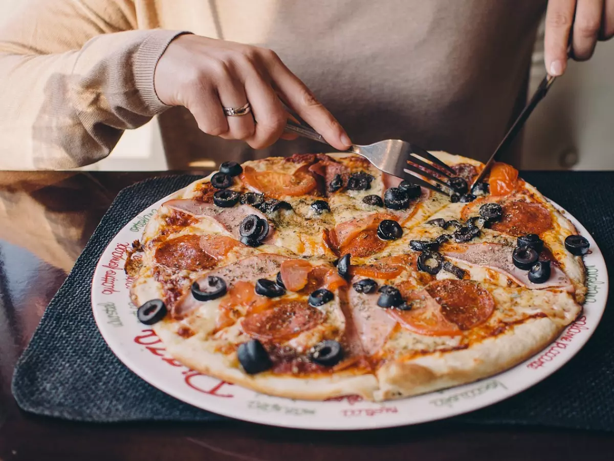 A close-up of a pizza with various toppings, including pepperoni, ham, olives, and cheese. The pizza is on a white plate and is being cut with a knife and fork.