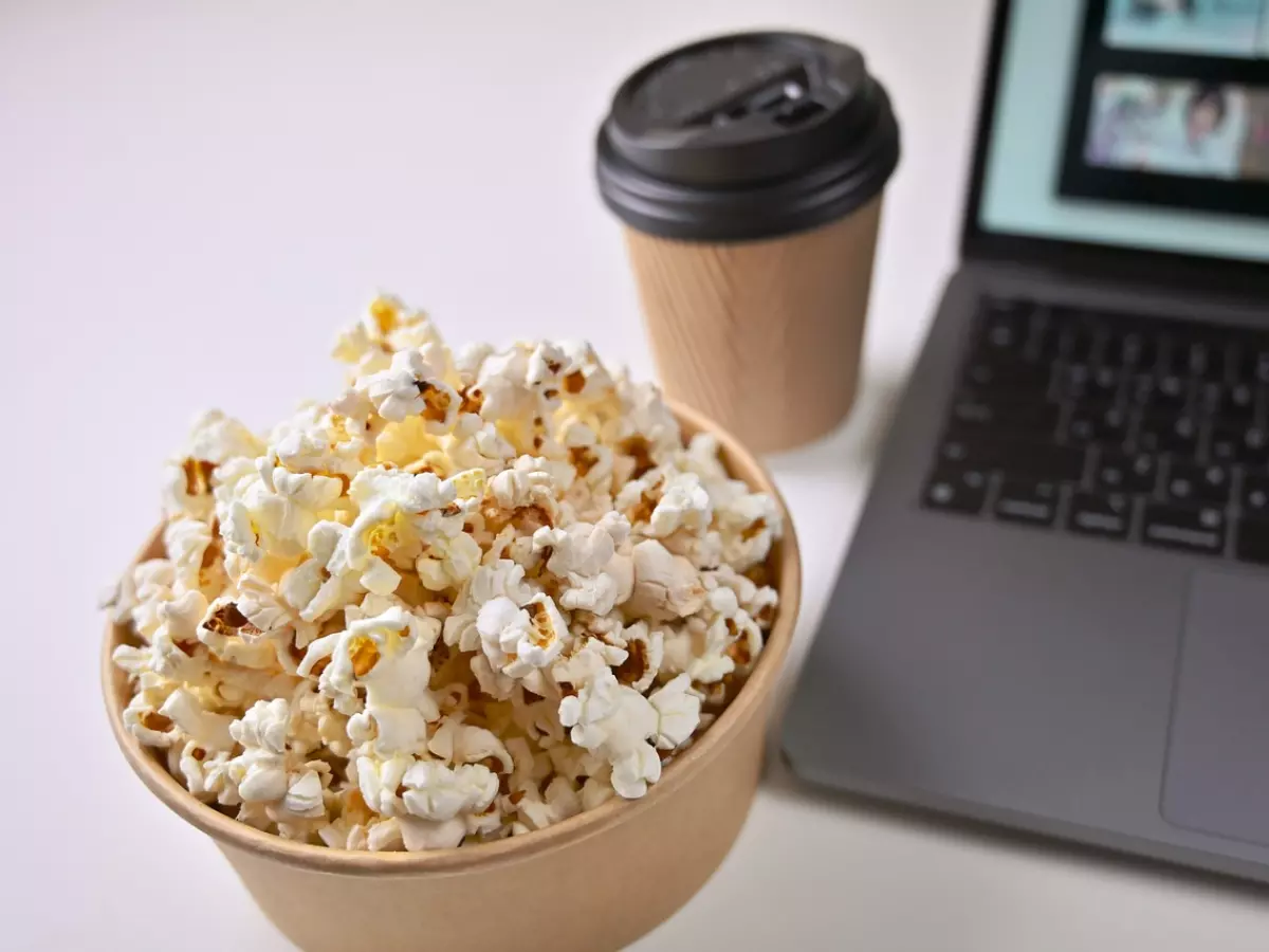 A laptop is on a white desk, with a bowl of popcorn in front of it. A cup of coffee is next to the popcorn.