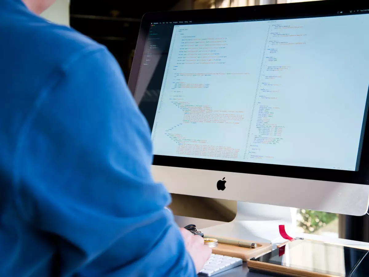 Person working on iMac with code on the screen, with a wireless keyboard, touchpad and mobile phone on the desk.