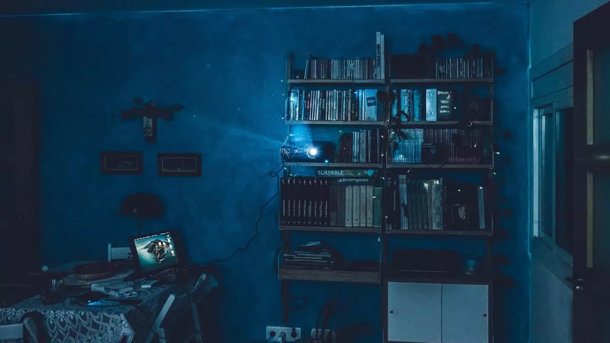 A projector is projecting on a wall in a living room. It is plugged to a laptop and pointed towards a bookshelf.