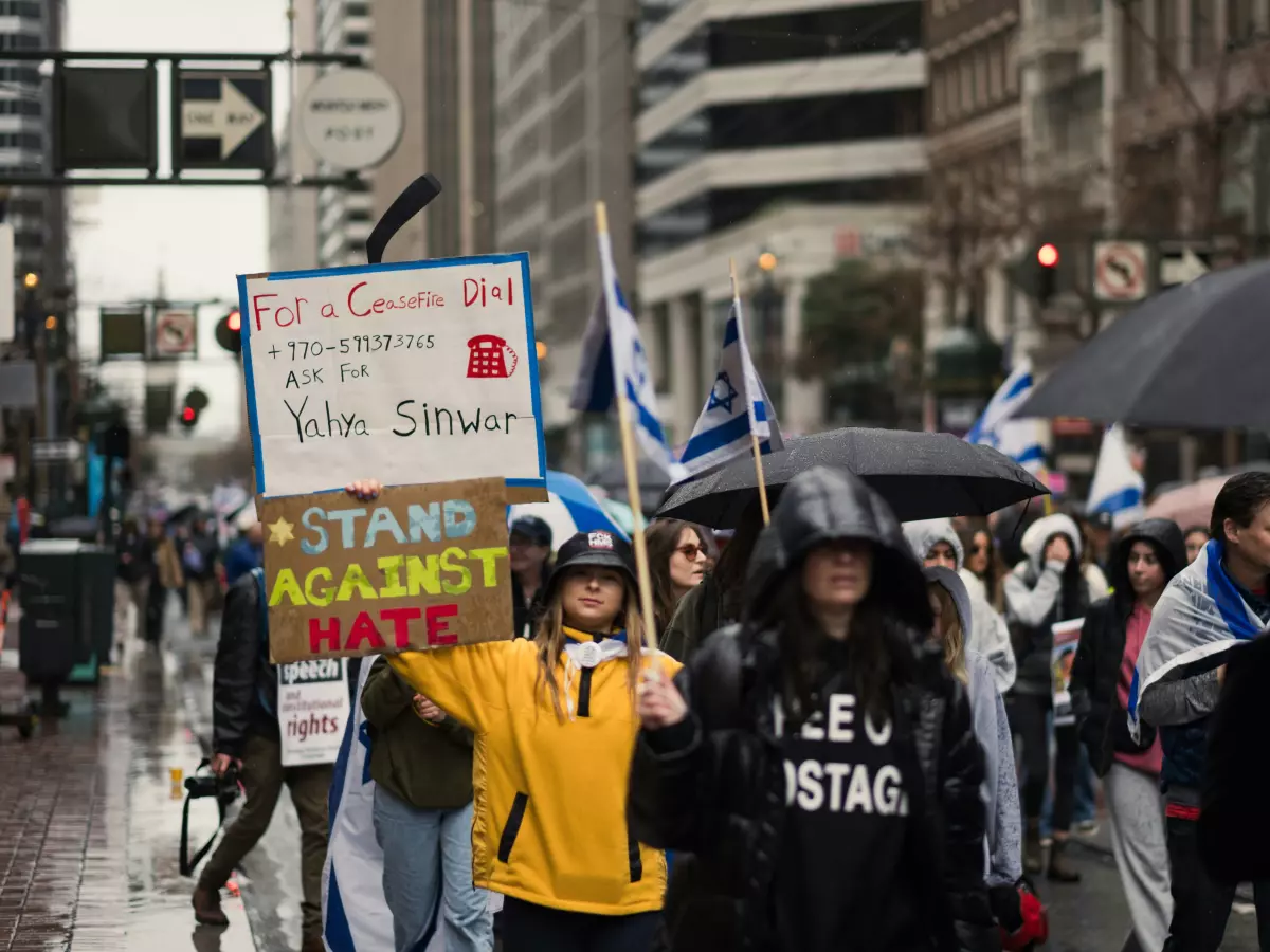 A group of people are protesting in the street. Some are holding signs that say "Stand Against Hate" and "I Stand Against Hate & Antisemitism".