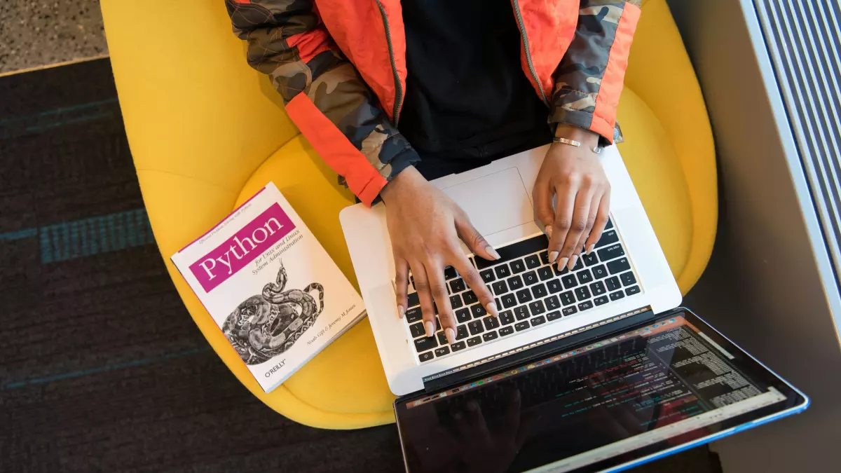 A person typing on a laptop with a book on the side.