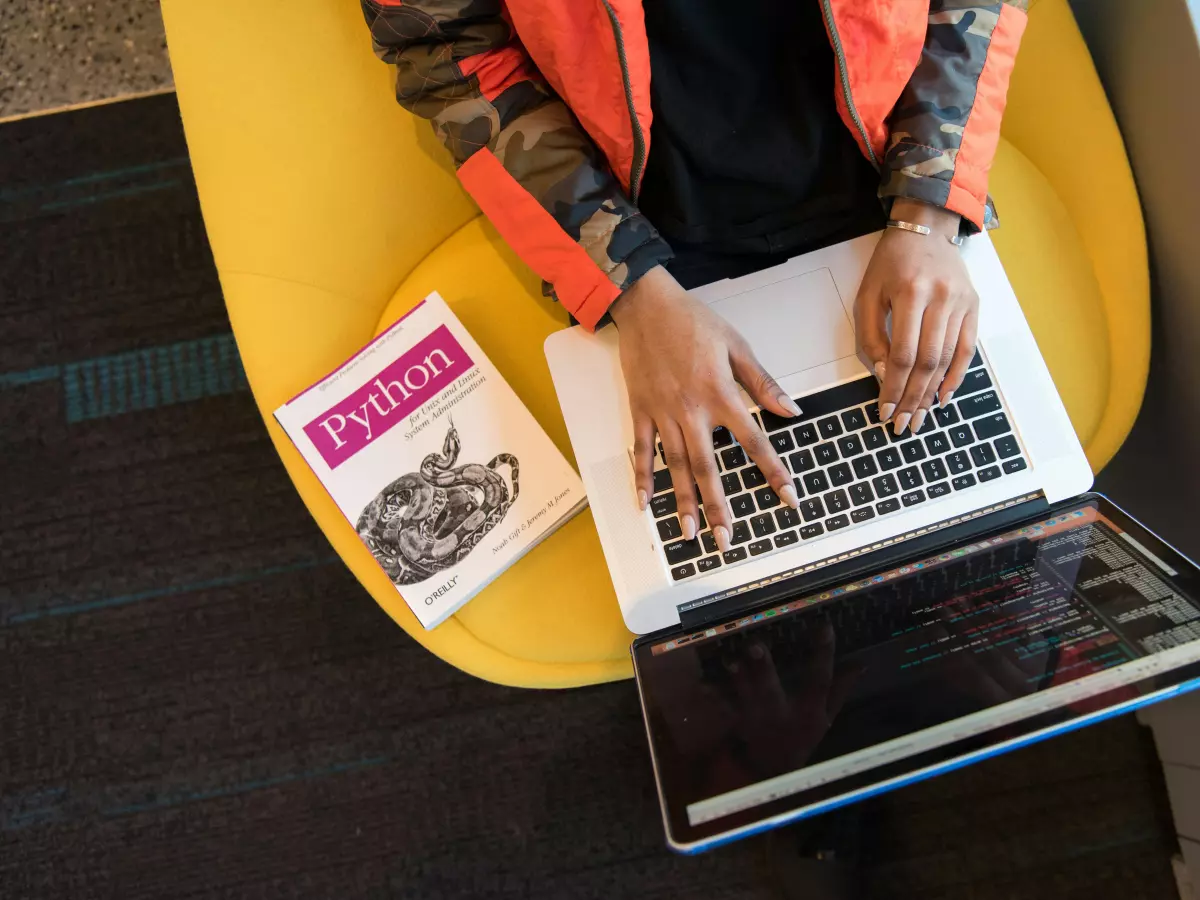 A person typing on a laptop with a book on the side.