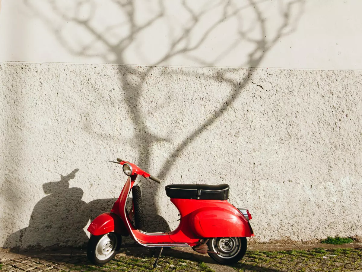 A red Vespa scooter parked in front of a white wall with the shadow of a tree behind it.