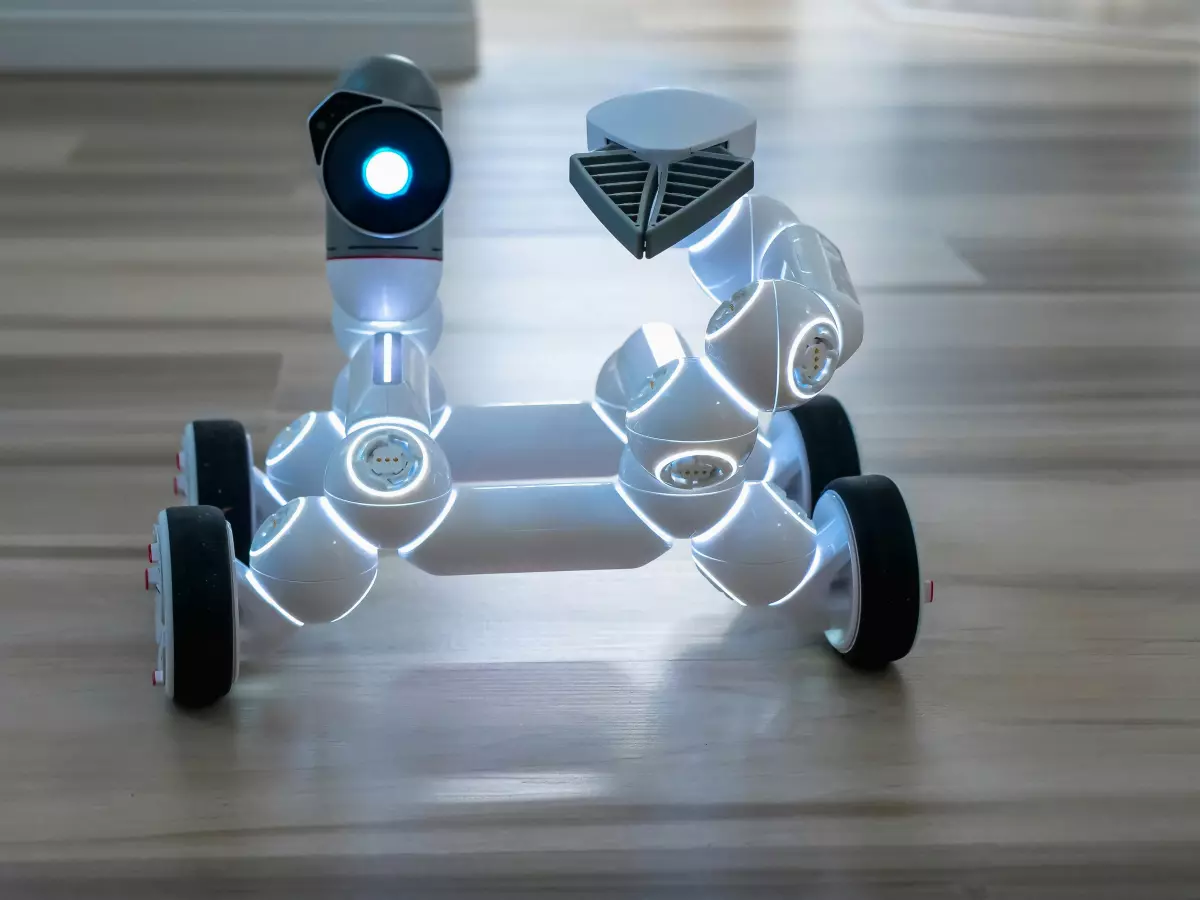 A white robot with black wheels on a wooden floor, the robot has visible sensors on its head and is in a slightly tilted position.