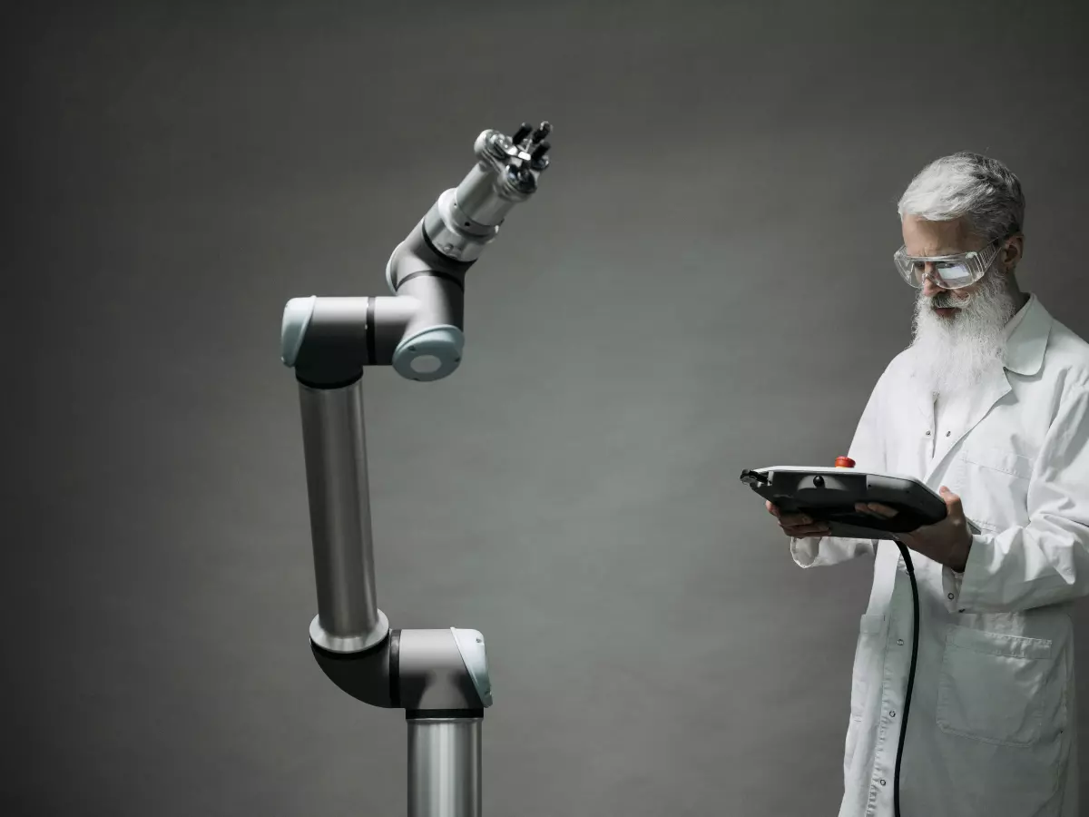 An older man with a white beard, wearing a white lab coat and glasses, uses a tablet computer while standing next to a robotic arm. The arm is in the background, reaching up and holding a small object.