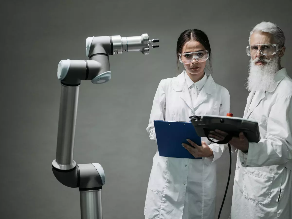 Two scientists in white coats stand in front of a robotic arm, they are looking at a device in the scientist's hand.