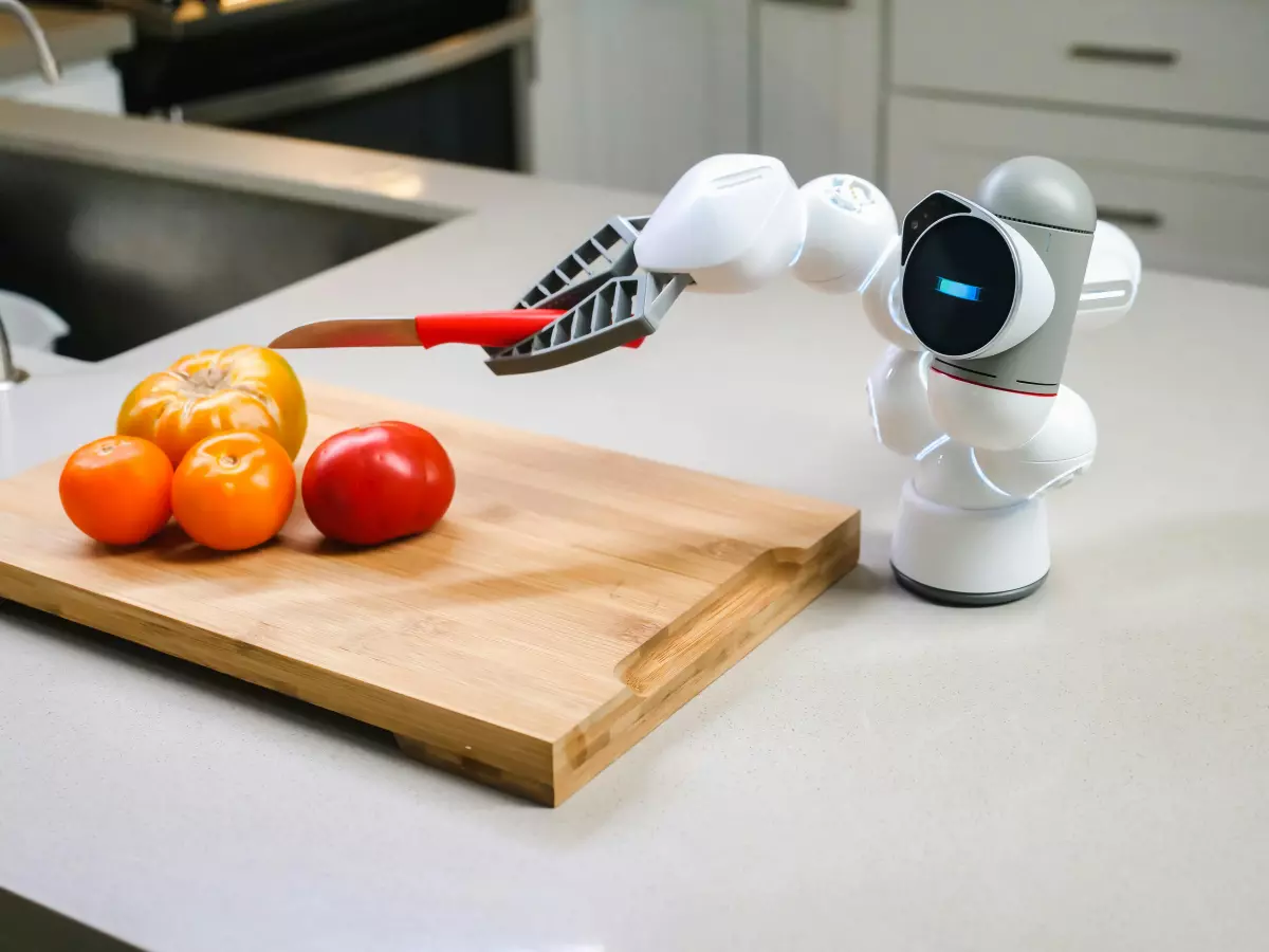 A white robot arm with a knife cuts a tomato on a wooden cutting board.