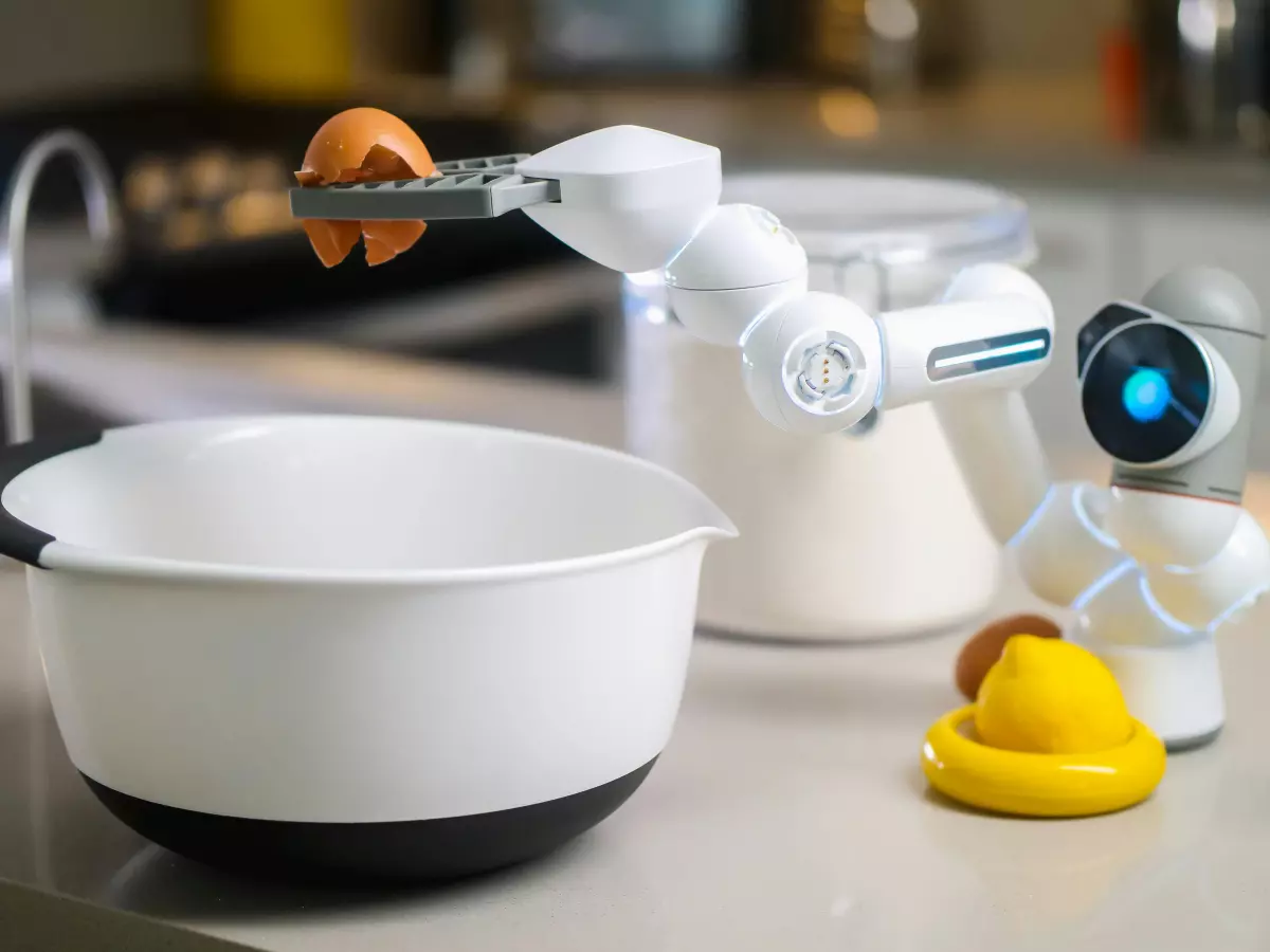 A white humanoid robot arm is picking up a brown egg with its robotic pincers. The robot is standing in a kitchen, with a bowl, a container of flour, and other kitchen items in the background.