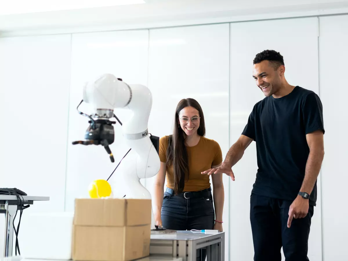 Two people look intently at a robotic arm moving a yellow object to a cardboard box.